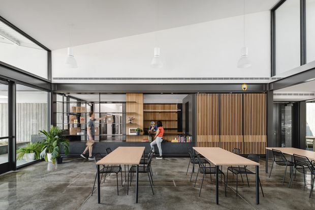 A bright, open-plan commercial interior featuring a communal dining and breakout area with multiple timber tables and black metal chairs. The space includes a dark service counter and kitchenette, extensive black-framed glazing, a large vertical timber slat feature wall, exposed polished concrete flooring, indoor planting, and suspended white pendant lights. A glazed meeting or work area is visible behind the counter.