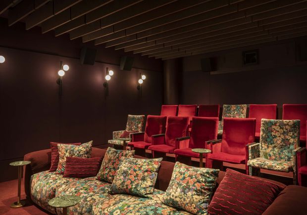 A dark, intimate home cinema or screening room with tiered rows of plush red upholstered theater seats, a floral-patterned sofa and armchairs in the foreground, small side tables, wall-mounted globe lights, and a ceiling lined with exposed linear timber battens. The rich red, brown, and patterned upholstery creates a theatrical, cozy atmosphere.