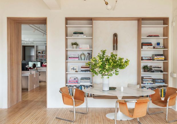 Bright, refined dining room with a round dining table and orange leather chairs, set against a wall of built-in timber-framed shelving with books and decorative objects. A central plastered feature panel and floral arrangement create a focal point. Timber flooring, warm neutral walls, and a doorway reveal an adjacent study or office.