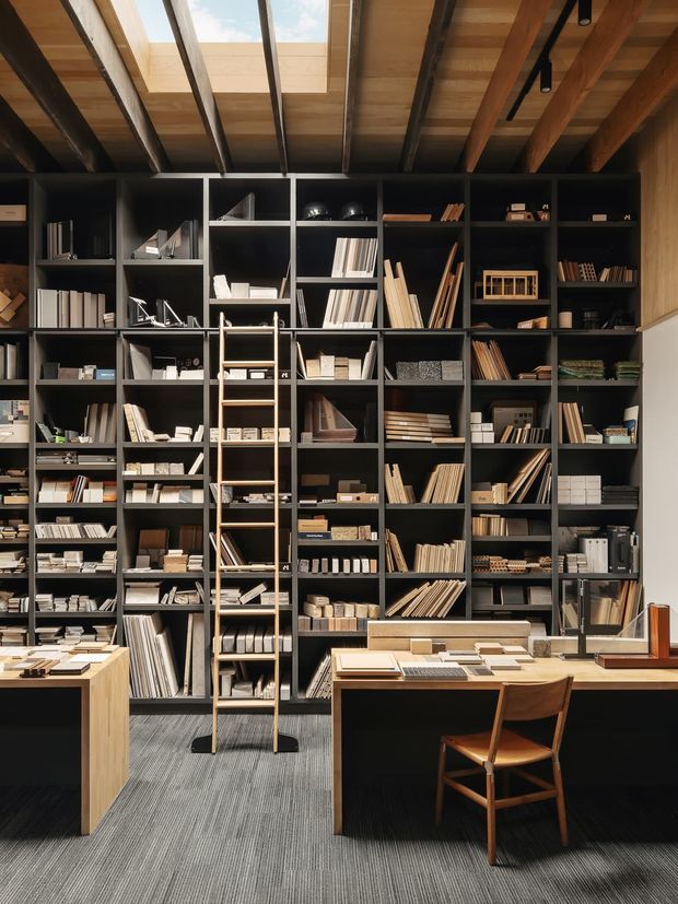 A refined interior workspace or materials library dominated by a full-height wall of dark built-in shelving filled with books, samples, drawings and design materials. A timber rolling ladder reaches the upper shelves, while timber desks with displayed material samples occupy the foreground. The space has exposed timber ceiling beams, a large skylight, dark joinery, and a subdued professional palette. The image appears to be a design studio, architectural materials library, or showroom workspace.