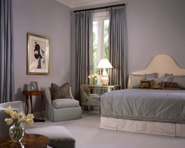Elegant, softly lit bedroom with pale lavender-grey walls, floor-length grey curtains, ornate crown moulding, and a tall window. The room features a large upholstered bed with a curved cream headboard and layered grey and patterned cushions, a mirrored bedside table with a table lamp, an upholstered armchair, a small dark wood side table, framed artwork, and a glass vase of flowers. The overall design is refined and traditional with a muted, luxurious palette.