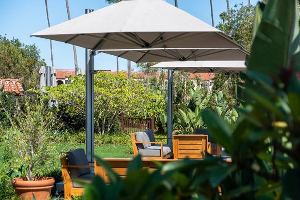 A landscaped outdoor seating area with timber lounge furniture beneath large square shade umbrellas. Dense tropical planting surrounds a lawn and timber privacy fence, with terracotta tiled roofs visible in the background. The scene is sunlit and resort-like.