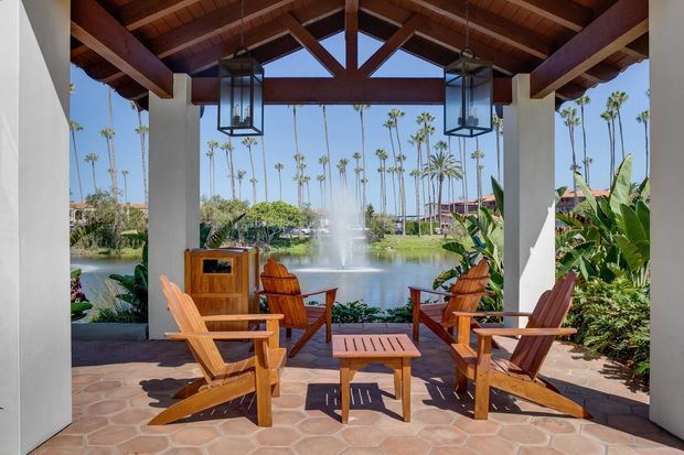 A covered outdoor lounge pavilion overlooking a landscaped lake, with timber Adirondack-style chairs and a low table arranged on a terracotta-toned irregular tile patio. White structural columns support a warm timber pitched roof with exposed beams, decorative trusses, and hanging lanterns. Tropical planting and tall palms surround the waterfront setting.