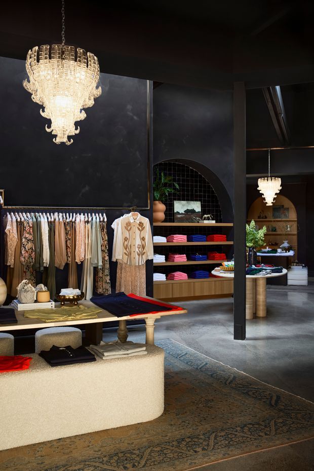 A moody, sophisticated clothing boutique interior with dark charcoal walls and ceiling, warm timber display shelving, clothing racks, folded garments, upholstered display benches, and ornate crystal chandeliers. An arched opening and reflective dark tile backdrop add architectural character, while a patterned rug softens the polished concrete floor.