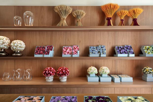 A warm, minimalist retail display wall with walnut-toned timber shelving. The shelves present neatly arranged floral bouquets and boxed flower arrangements in varied colours, alongside clear glass vases and decorative vessels.