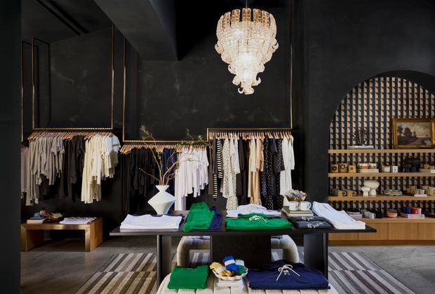 A dramatic, high-end clothing boutique interior with charcoal-black walls, open garment racks, neatly folded apparel on central display tables, and a decorative arched shelving niche at the rear. A large crystal chandelier provides a luxurious focal point, while timber cabinetry and patterned rugs add warmth and texture.