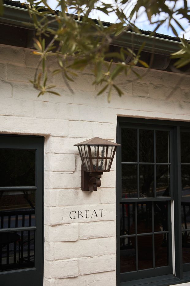 Close-up exterior view of a white-painted brick facade with dark multi-pane framed windows and a traditional wall-mounted lantern. Olive branches partially frame the upper portion of the image, and signage reading “THE GREAT.” is visible on the wall.