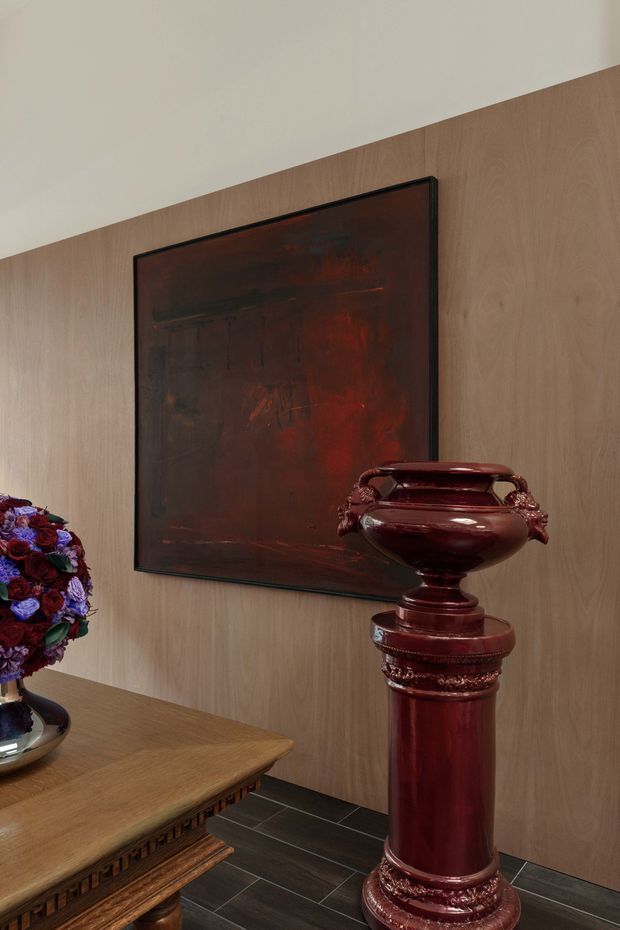 A refined interior vignette featuring a warm timber wall panel, a large dark abstract artwork in a black frame, and an ornate deep burgundy ceramic urn displayed on a matching pedestal. Part of a carved timber table and a floral arrangement are visible at the left, with dark tiled flooring below. The composition is richly coloured and decorative, with a strong traditional and luxurious character.