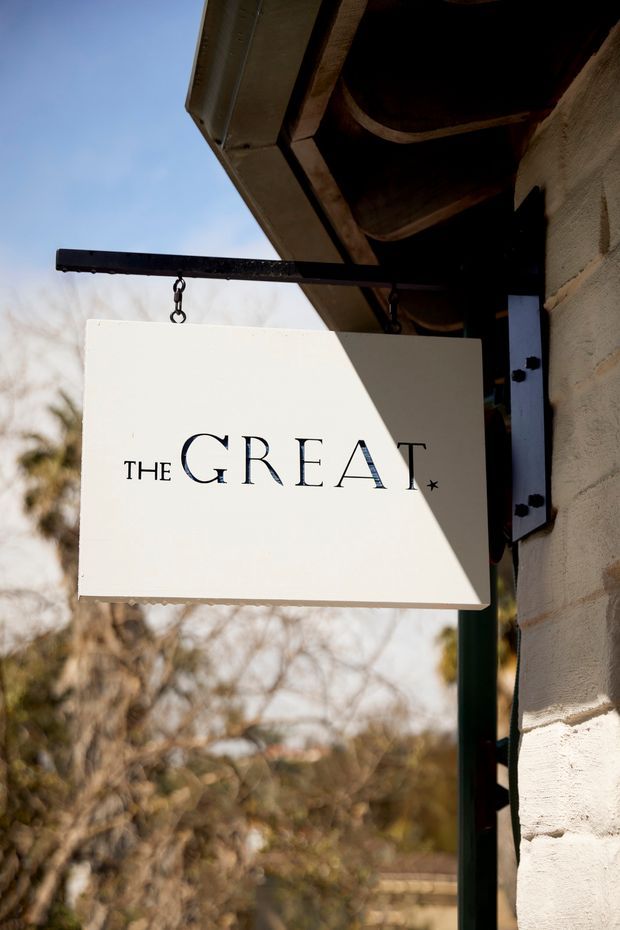 Close-up exterior photograph of a hanging rectangular sign reading “THE GREAT” beneath a timber roof eave. The sign is suspended from a dark metal bracket attached to a textured light-coloured masonry wall, with trees and sky softly blurred in the background.