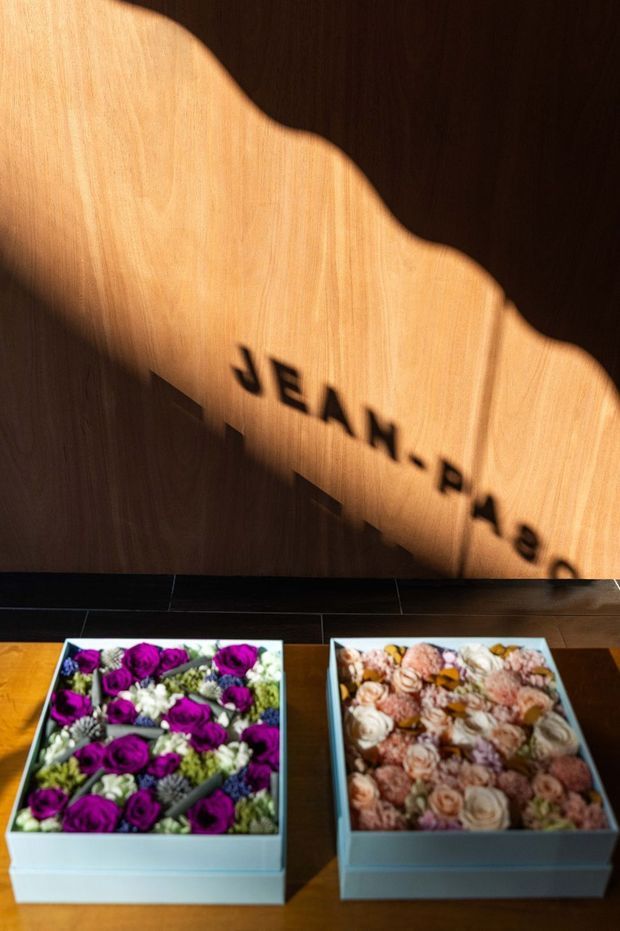 A warmly lit decorative display featuring two open rectangular boxes of arranged flowers on a dark timber surface. A timber wall behind them shows dramatic scalloped shadowing and partially visible lettering reading “JEAN PAUL…”, creating a boutique retail or reception-like atmosphere.
