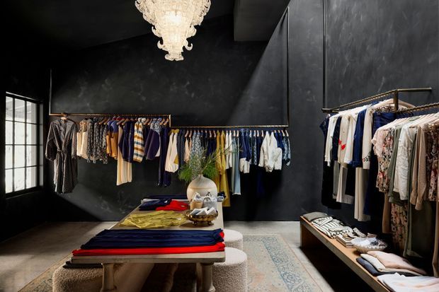 A dark, luxurious fashion retail showroom with black textured walls, clothing racks lining the walls, a central display table with folded garments, upholstered stools, a patterned rug, and an ornate crystal chandelier. Large industrial-style window panes bring in natural light from the left.