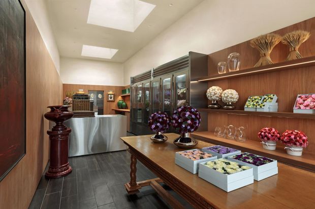 A refined floral display and retail preparation space with dark timber wall panelling, built-in open shelving, glass-fronted cabinetry, a central wooden display table, and a stainless-steel work counter. Fresh floral arrangements and boxed flowers are arranged throughout, with skylights providing bright overhead illumination.
