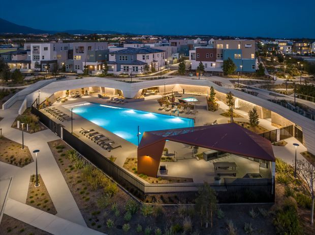 Elevated dusk view of a contemporary residential development centred around a brightly lit swimming pool and landscaped communal outdoor recreation area. Curved paved pathways, lounge seating, shade structures, low planting, and surrounding multi-storey apartment or townhouse buildings are visible, with hills and a suburban settlement in the distance.