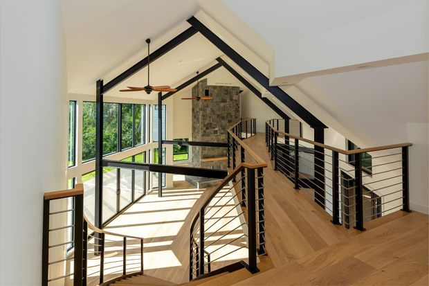 Bright multi-level interior with a dramatic vaulted ceiling and exposed black structural beams. A timber-floored landing and bridge are enclosed by black metal cable railings, overlooking a sunlit double-height glazed space. A stone-clad feature wall and ceiling fans add visual interest, while abundant glazing brings in greenery and daylight.