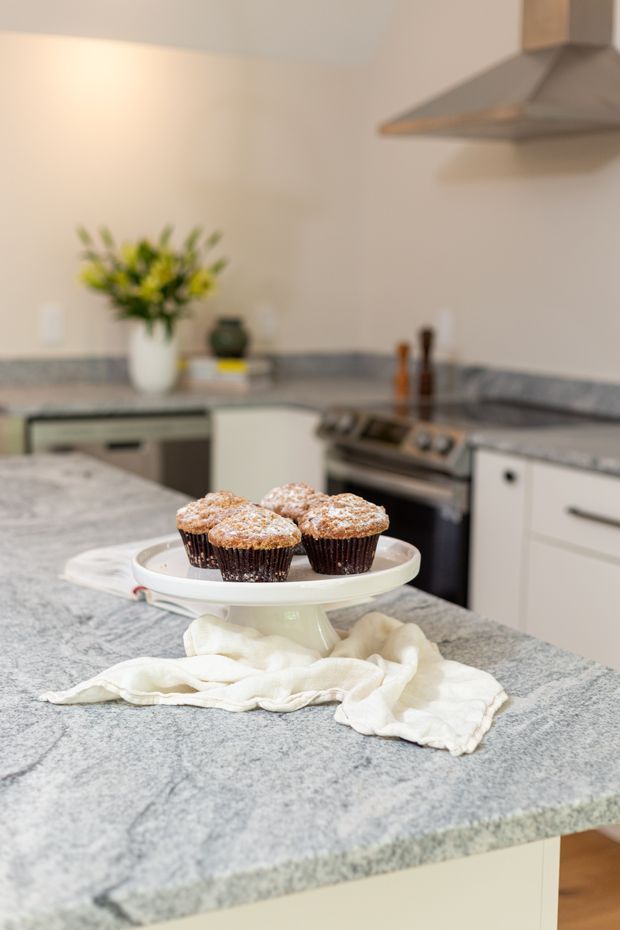 A softly lit kitchen interior with a large grey stone island benchtop in the foreground. A white pedestal cake stand holding muffins sits on the island beside a folded white cloth. In the background are pale cabinetry, a stainless-steel rangehood and oven, grey stone perimeter benchtops, and a small floral arrangement.