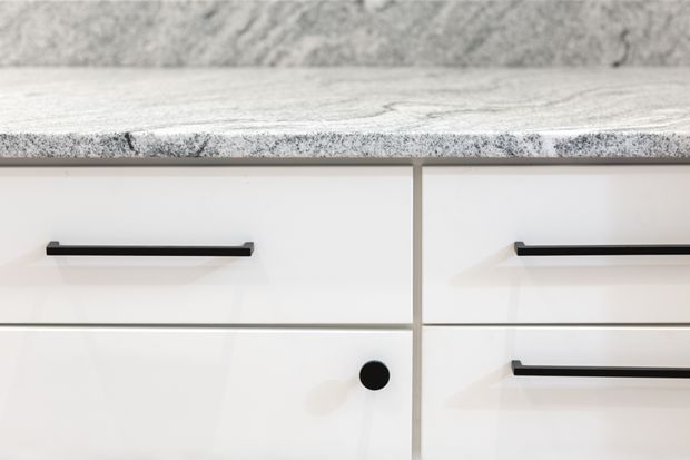 Close-up detail of white built-in cabinetry with slim black bar handles and a black round knob. A light grey speckled stone benchtop or surface sits above the drawers, with a textured grey wall or splashback behind it. The image focuses on clean, contemporary joinery and contrasting hardware.