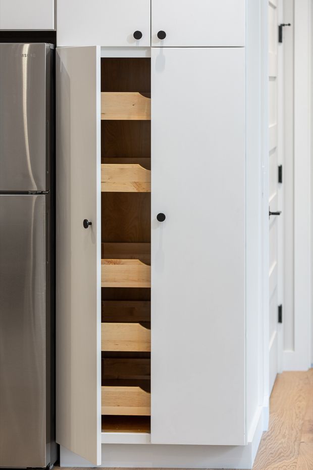 Close-up interior view of a tall white built-in pantry cabinet beside a stainless-steel refrigerator. The pantry door is partially open, revealing multiple light timber pull-out shelves and a dark timber interior. White cabinetry continues above and beside the pantry, with black round knobs. A light timber floor and a white hallway with black hardware are visible to the right.
