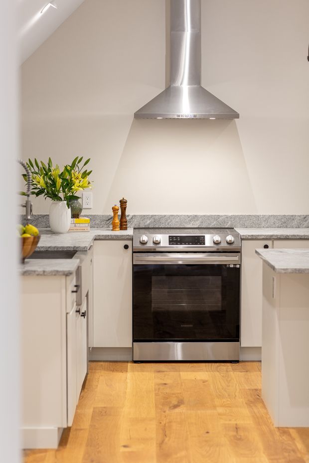 A compact, bright kitchen with a freestanding stainless-steel oven and rangehood centered against a light painted wall. Pale cabinetry with dark hardware, grey stone-look benchtops and splashback, and warm timber flooring create a clean, contemporary fit-out. A small island or peninsula frames the foreground, with minimal decorative styling and potted greenery.