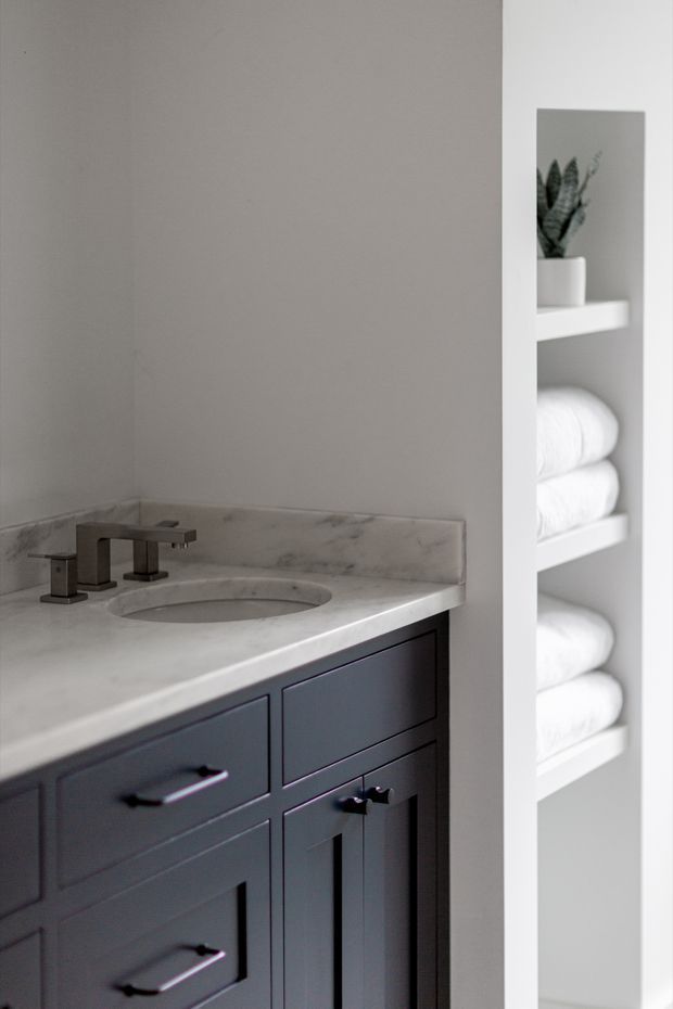 A refined bathroom vanity with a dark painted timber-style cabinet, pale marble countertop and integrated oval basin. Brushed metal tapware sits against a simple light wall, while open recessed shelving displays neatly folded white towels and a small potted plant.