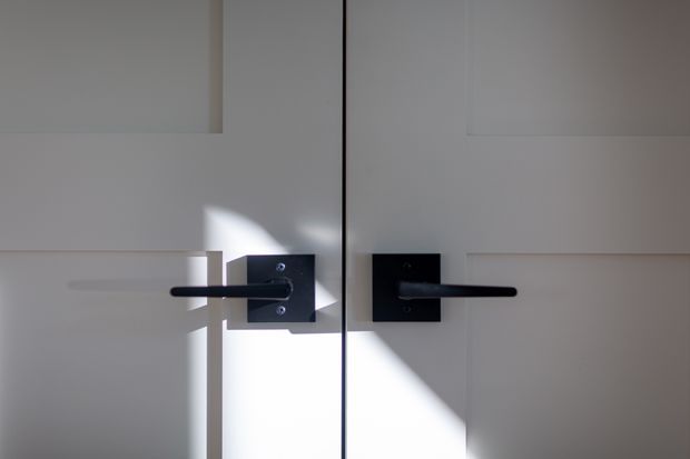 Close-up architectural detail of paired white panelled doors with matte black square backplates and horizontal lever handles. A narrow central gap and strong diagonal sunlight create a high-contrast, minimalist composition.
