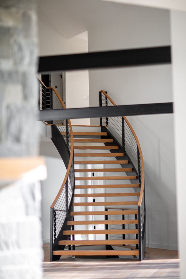 A contemporary interior stairway with open-riser timber treads, dark metal stringers and horizontal wire railings, plus warm timber handrails. The staircase curves gently upward through a bright, white-painted multi-level space, with a landing visible above and timber flooring at the base.