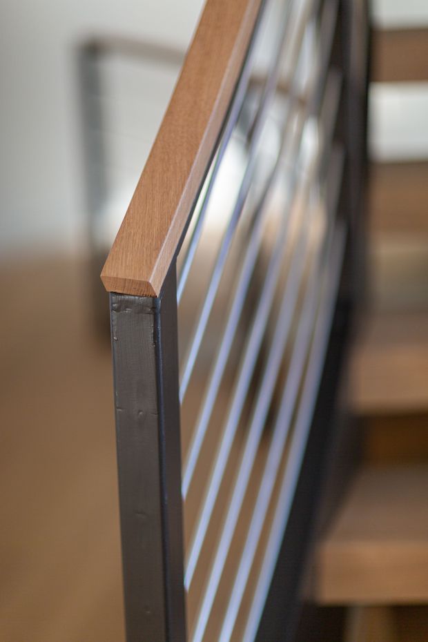 Close-up detail of a contemporary stair balustrade featuring a warm timber handrail mounted on a dark metal frame, with multiple slender vertical metal balusters. Timber stair treads are softly visible in the background.