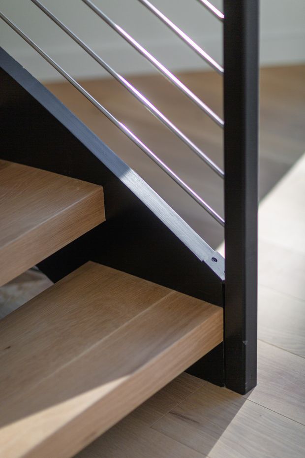 Close-up architectural photograph of a contemporary open-riser staircase. The stair treads have a light natural timber finish, supported by a dark structural stringer and post, with slim horizontal metal balusters forming the railing. Light timber flooring continues around the stair.
