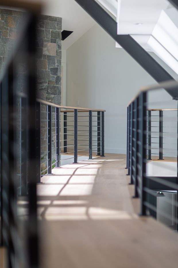 A bright contemporary interior landing or upper-level hallway with an open stair void. The space features dark metal balustrades with horizontal cable rails and timber handrails, a pale smooth floor, white walls, high clerestory-like glazing, and a prominent rough stacked-stone feature wall. Strong sunlight casts geometric shadows across the floor and emphasizes the open, airy volume.