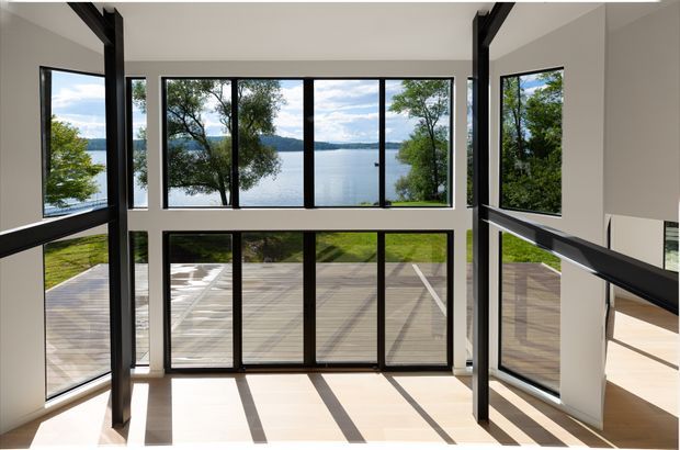 A bright double-height interior with expansive black-framed floor-to-ceiling glazing overlooking a timber deck, lawn, trees, and a calm body of water. Strong linear shadows fall across the pale floor, and dark structural posts frame the open void.