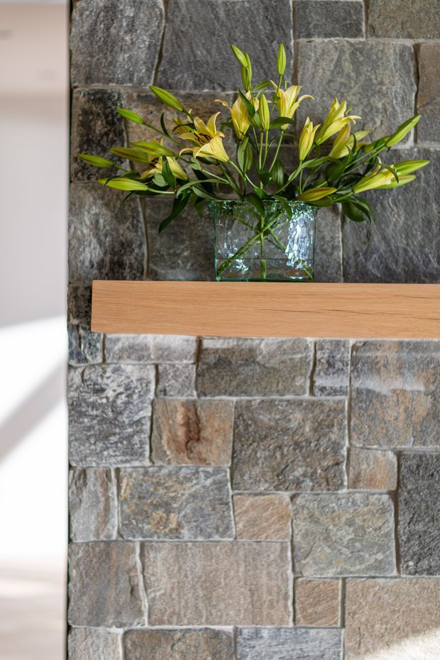 Close-up interior detail of a rustic stone-clad feature wall with a slim floating timber shelf and a clear glass vase of lilies. Natural light enters from the side, highlighting the varied grey, brown, and tan tones of the masonry.
