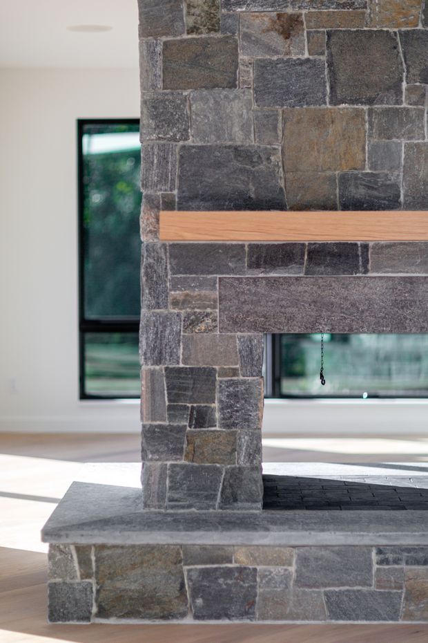 Close-up interior view of a substantial fireplace constructed from irregular dark grey and brown natural stone masonry, with a timber mantel shelf and dark stone hearth. Large black-framed windows behind the fireplace bring in abundant daylight and views of greenery.