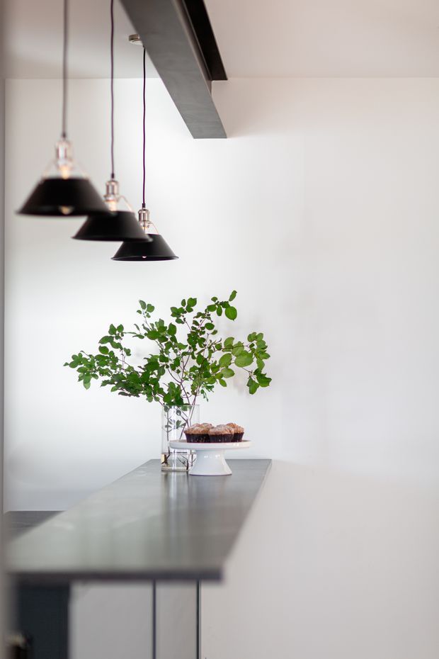 Minimal, bright interior detail featuring a long grey stone or concrete kitchen island/benchtop, three black pendant lights with exposed cords, and a simple floral arrangement with baked goods. White walls and ceiling create a clean, restrained composition.
