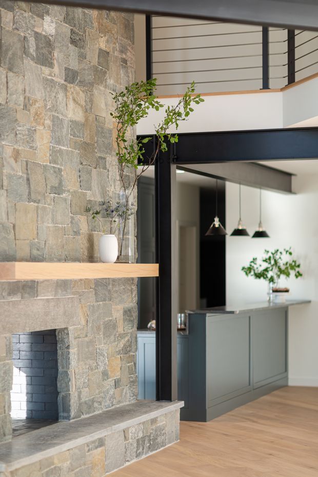 A refined open-plan interior featuring a substantial natural stone fireplace wall with a simple timber mantel, black steel structural framing, and an upper-level balustrade. Beyond the opening is a contemporary kitchen with dark grey cabinetry, black pendant lights, and timber flooring. Potted branches and minimalist detailing soften the industrial-architectural composition.