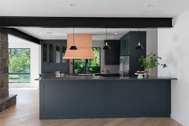 A contemporary kitchen viewed from the living area, featuring a large dark painted island with a stone benchtop, matching dark cabinetry, integrated stainless-steel appliances, and three pendant lights. A distinctive copper-toned rangehood is suspended above the cooktop. Timber flooring, a black exposed structural beam, and large windows add warmth and natural light.