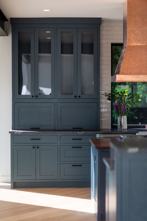 A sophisticated kitchen with floor-to-ceiling dark blue-grey shaker cabinetry, glazed upper doors, integrated drawers and lower cupboards, and a dark stone benchtop. A pale brick tile splashback, copper-toned rangehood, timber flooring, and a kitchen island are visible, with strong sunlight entering through a window beside the cabinetry.