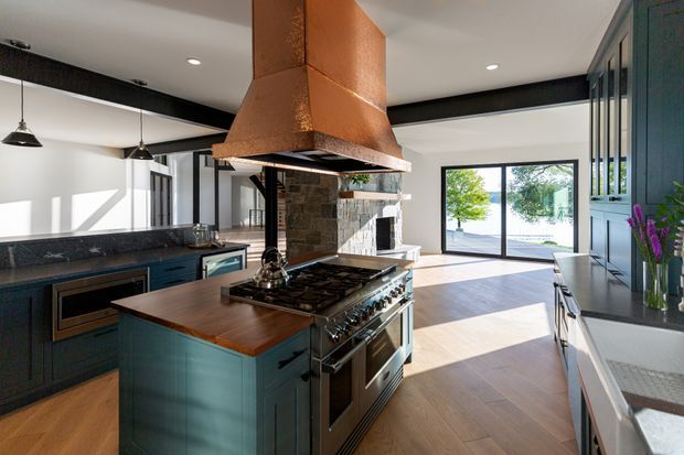 Open-plan contemporary kitchen with a large central cooking island, dark teal cabinetry, a timber benchtop, dark stone perimeter counters, stainless-steel range cooker, and a prominent copper-clad extraction hood. A stone fireplace feature and glazed living area are visible beyond the kitchen.