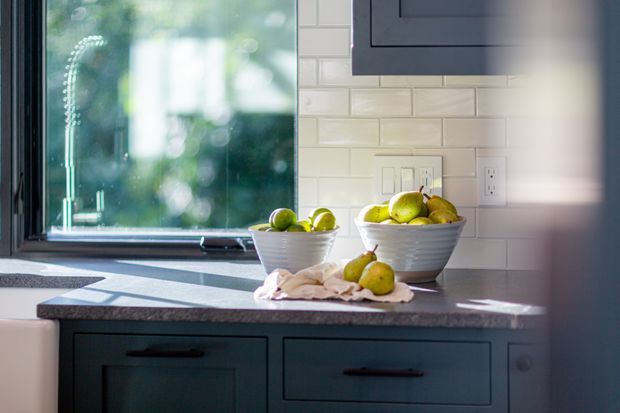 Close-up view of a kitchen benchtop beneath a window, featuring dark blue-grey cabinetry, a grey stone-look counter, white subway tile splashback, electrical outlets, and bowls of green fruit. Natural light enters through the black-framed window.