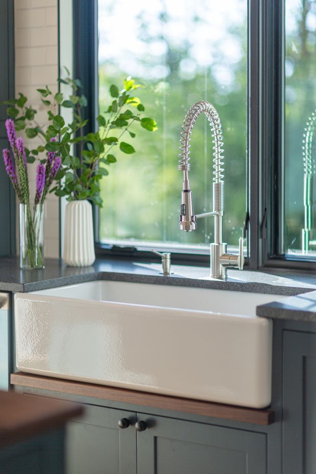 Close-up of a farmhouse-style kitchen sink area featuring a large white apron-front fireclay sink, dark grey stone benchtops, dark painted cabinetry, and a polished chrome spring-coil gooseneck faucet. Tall windows provide abundant natural light, with plants and flowers arranged beside the sink.