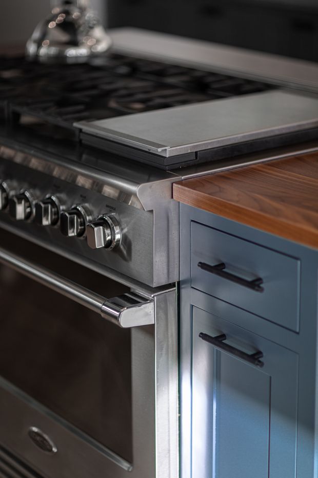 Close-up detail of a dark, high-end kitchen range cooker beside blue built-in cabinetry with black handles and a warm timber benchtop. The cooker has a stainless-steel front, heavy control knobs, oven handle, and a dark gas cooktop with a pale rectangular griddle or cover.