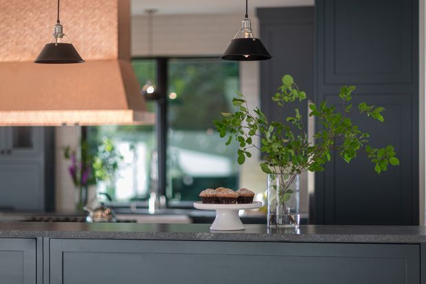 A refined kitchen interior viewed across a dark painted island or peninsula. The space features a large warm copper-toned range hood, dark charcoal cabinetry with framed panel detailing, a grey stone benchtop, black pendant lights, and a reflective splashback or window behind the work area. A vase of leafy branches and a cake stand provide restrained decorative accents.