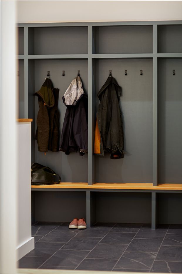 A built-in mudroom or entry storage wall with dark grey painted cubby joinery, open coat compartments, hooks, timber bench seating, lower shoe storage, and dark tiled flooring. Jackets, shoes, and a bag are visible within the storage units.