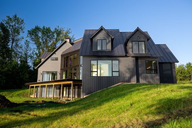Exterior view of a contemporary multi-level house on a sloping, grassy site. The home features dark vertical cladding, standing-seam metal gable roofs, projecting dormer windows, extensive glazing, and a partially elevated lower level surrounded by mature trees.