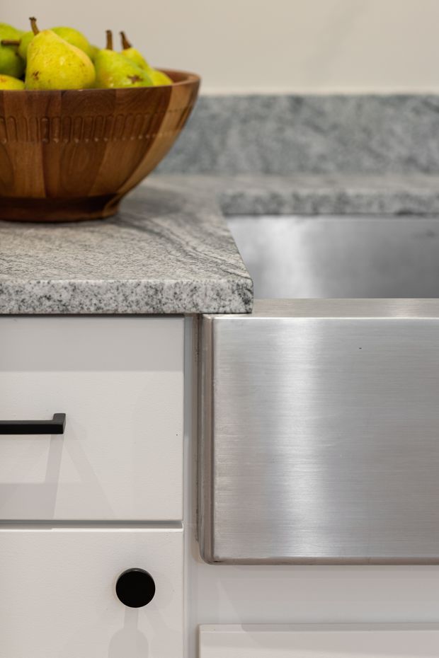 Close-up detail of a contemporary kitchen, showing grey granite benchtops, white cabinetry with black hardware, and a large brushed stainless-steel drawer or appliance front. A wooden bowl of pears sits on the benchtop beside a stainless-steel sink.