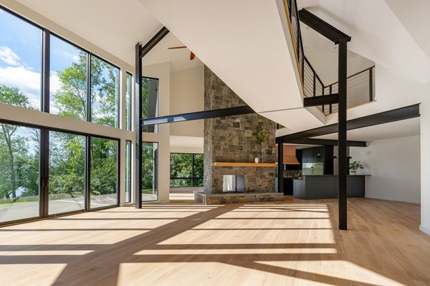 A bright, expansive double-height open-plan interior with floor-to-ceiling glazing, pale timber flooring, a tall rough stone fireplace feature wall, and exposed black steel structural posts and beams. An upper-level mezzanine with black metal balustrading overlooks the living area, while a compact dark kitchen is visible at the rear. Strong sunlight creates long geometric shadows across the floor.