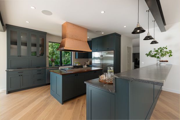 A spacious, refined kitchen with dark blue-grey shaker-style cabinetry, two substantial islands, and a striking copper rangehood canopy. Pale timber flooring, white walls and ceiling, grey stone benchtops, black pendant lights, and exposed dark ceiling beams create a warm contemporary-traditional interior. Glazing above the rear benchtop brings in greenery and natural light.