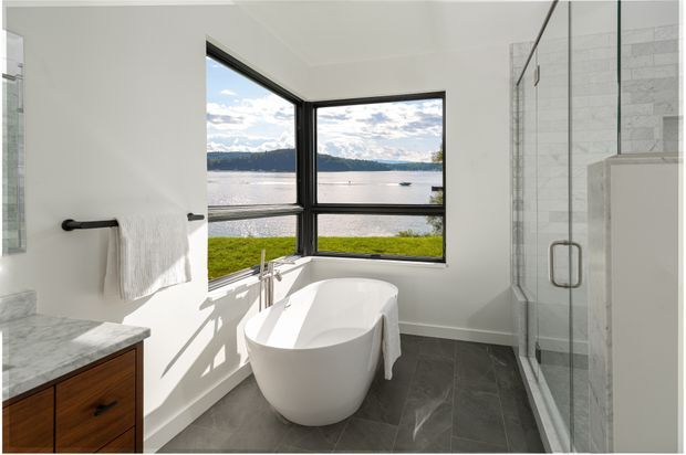 Bright contemporary bathroom with a freestanding white bathtub positioned beside large corner windows overlooking a lake and hills. The room includes a glass-enclosed shower, dark grey floor tiles, white wall finishes, black window and hardware details, and a timber vanity with a pale stone benchtop.