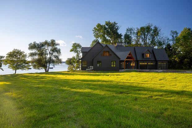 A dark-clad, multi-gabled house set in a broad green lawn beside a calm body of water. The home features steep pitched roofs with dark metal roofing, timber-framed gable accents, warm illuminated windows, and an attached garage. Mature trees surround the property under a clear blue sky.