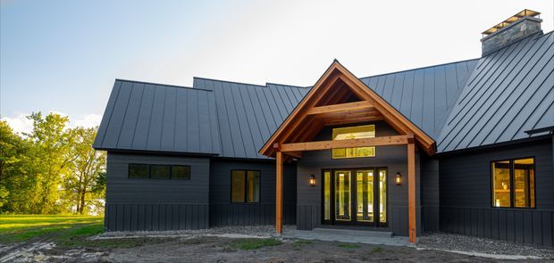 Exterior view of a dark-clad, single-storey house with a steep metal gabled roof and prominent timber-framed gable entry porch. The facade includes narrow horizontal windows, glazed double entry doors, warm timber accents, and a masonry chimney, set within a leafy landscape.