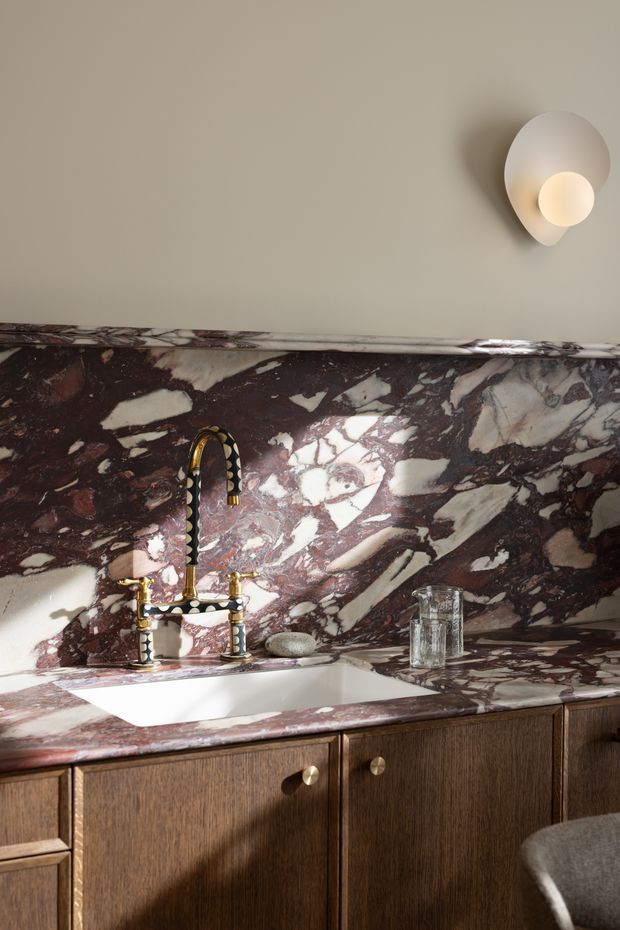 Close-up of a refined bathroom vanity featuring a dramatic burgundy and cream veined stone backsplash and benchtop, an integrated white rectangular basin, ornate brass tapware, and dark timber cabinetry with round brass knobs. A sculptural wall sconce provides warm illumination above the vanity.