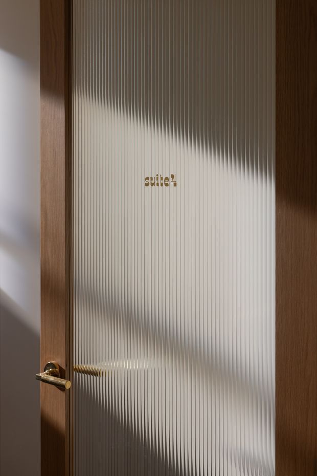 Close-up architectural view of a timber-framed interior door with vertically ribbed/frosted glass. A brass or gold-toned lever handle is visible, along with a small gold 'suite 2' sign on the glass. The textured glazing obscures the room beyond, while warm timber and neutral wall surfaces create a refined, softly lit entrance detail.